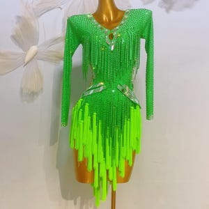 May include: A green sequined dance dress with long sleeves and a fringe skirt. The dress is decorated with rhinestones and has a V-neckline.