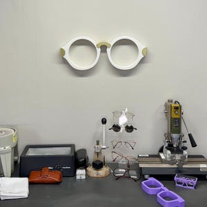 Op de afbeelding: Een werkruimte van een opticien met diverse brillen en gereedschappen. Een groot wit en goudkleurig brilmontuur is aan de muur bevestigd. Verschillende brillen worden getoond, samen met een lenssnijder en andere apparatuur.