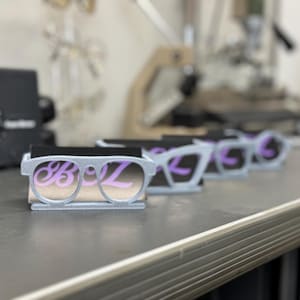May include: A row of gray eyeglasses-shaped business card holders with the letters "Boz" in a light purple color. The holders are on a gray surface, with a blurred background.