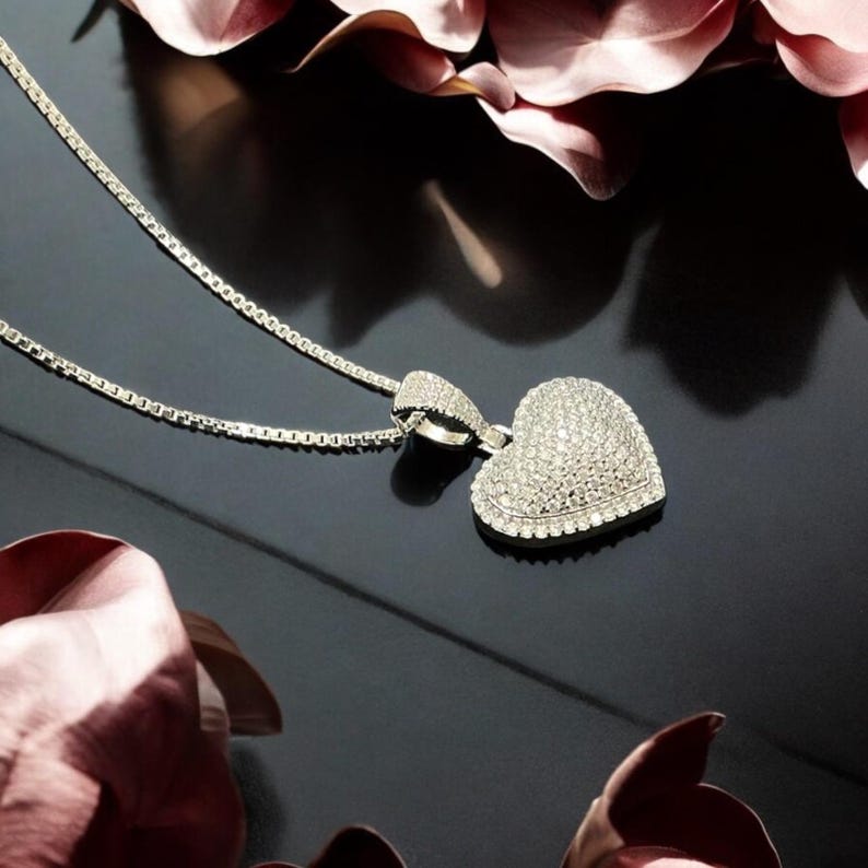 May include: A silver necklace with a heart-shaped pendant encrusted with sparkling crystals. The pendant is the focal point, reflecting light against a dark background. Pink petals frame the jewelry.