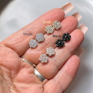 May include: Close-up of a hand displaying five pairs of flower-shaped earrings in gold, silver, rose gold, and black.