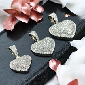 May include: Three heart-shaped pendants encrusted with small, sparkling stones. Each pendant has a silver-colored bail for a chain. The pendants are displayed on a dark, textured surface, with pink and white floral accents in the background.