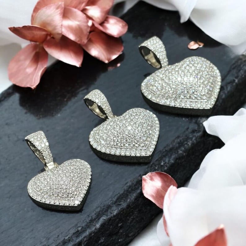 May include: Three heart-shaped pendants encrusted with small, sparkling stones. Each pendant has a silver-colored bail for a chain. The pendants are displayed on a dark, textured surface, with pink and white floral accents in the background.