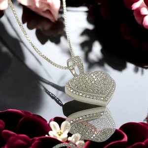 May include: A silver necklace with a heart-shaped pendant. The pendant is encrusted with small, sparkling stones, and the chain is a delicate silver. The heart is reflected on a shiny surface, surrounded by red petals and flowers.