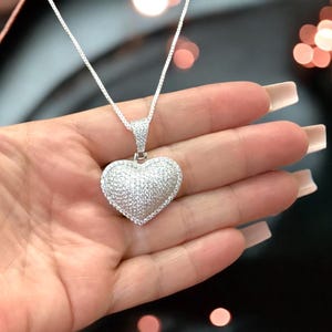 May include: A silver necklace featuring a heart-shaped pendant. The pendant and the bail are encrusted with small, sparkling stones. The necklace is displayed against a blurred background.