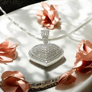 May include: A silver pendant necklace with a heart-shaped design, encrusted with numerous small, sparkling stones. The pendant is suspended from a delicate silver chain. The image is set against a white background with pink floral accents.