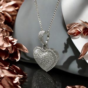 May include: A silver heart pendant necklace with a box chain. The heart-shaped pendant is encrusted with small, sparkling stones. The necklace is displayed against a gray background with rose-colored petals.
