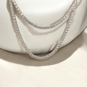 May include: Two silver-toned tennis necklaces with clear, sparkling stones. The necklaces are layered, with one resting slightly above the other. The jewelry is displayed against a white background, highlighting the brilliance of the stones.