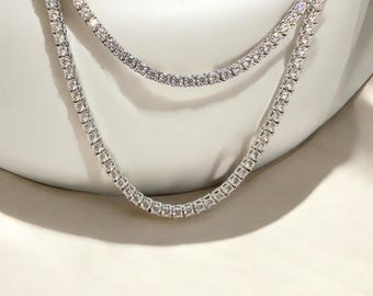 May include: Two silver-toned tennis necklaces with clear, sparkling stones. The necklaces are layered, with one resting slightly above the other. The jewelry is displayed against a white background, highlighting the brilliance of the stones.
