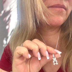 May include: A silver necklace featuring a small teddy bear charm. The bear is wearing a red heart and has sparkling accents. The necklace is being held by a hand with white-tipped nails.