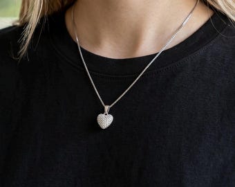 May include: A silver necklace with a heart-shaped pendant. The pendant is covered in small, sparkling stones. The necklace is worn against a black shirt, creating a contrast that highlights the jewelry.