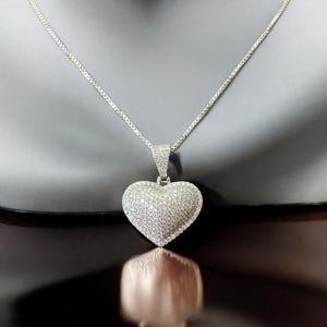 May include: A silver necklace featuring a heart-shaped pendant. The pendant and the bail are encrusted with small, sparkling stones. The chain is a delicate box chain, and the pendant is centered in the frame.