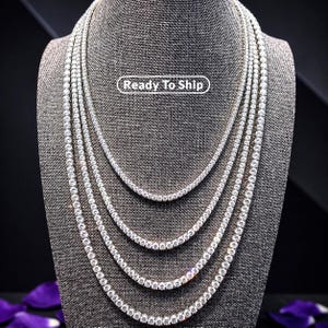 May include: Four layered silver-toned tennis necklaces featuring round clear stones set in a continuous chain design. The necklaces are displayed on a textured gray bust. A white rectangular label with the text Ready To Ship is centered.