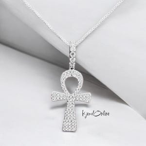 May include: A silver-toned ankh pendant necklace with a delicate chain. The ankh is encrusted with small, sparkling stones. The pendant is centered in the frame against a white background. The text "KjewlOnline" is in the lower right corner.