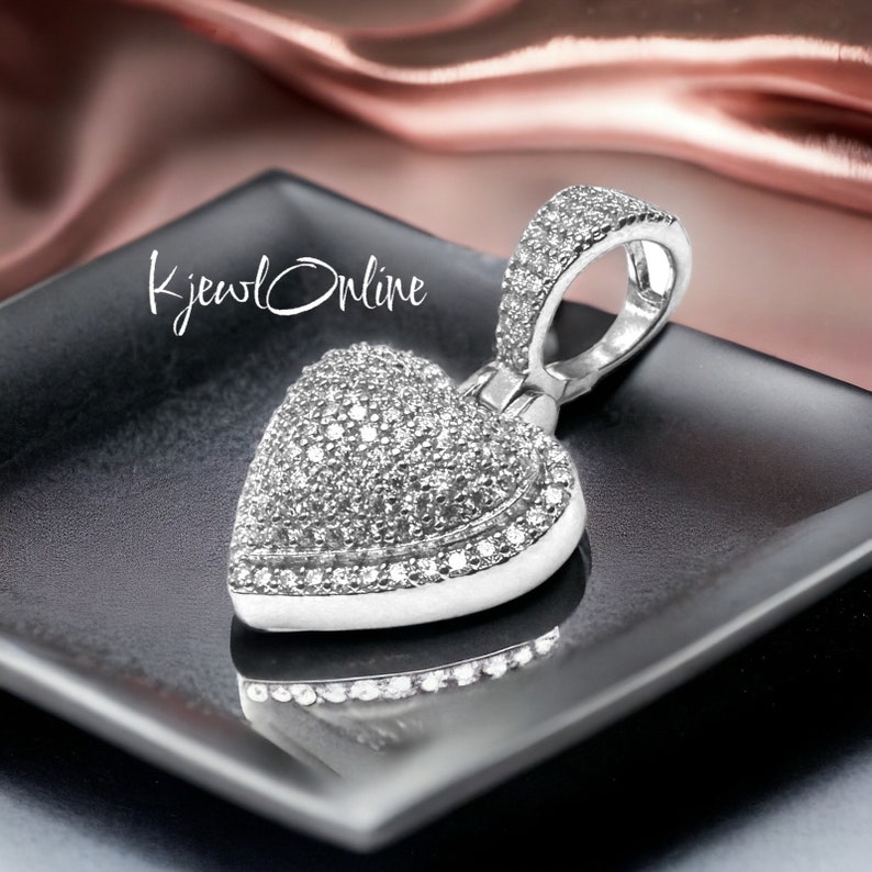 May include: A heart-shaped pendant with a pav&eacute; design, set with numerous small, sparkling stones. The pendant is silver-toned and rests on a black square surface. The text "KjewlOnline" is visible in the image.