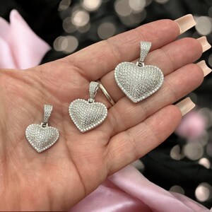 May include: Three silver heart pendants, each encrusted with small, sparkling stones. The pendants vary in size, with a bail at the top for a chain. The hearts are set against a blurred background with pink fabric.