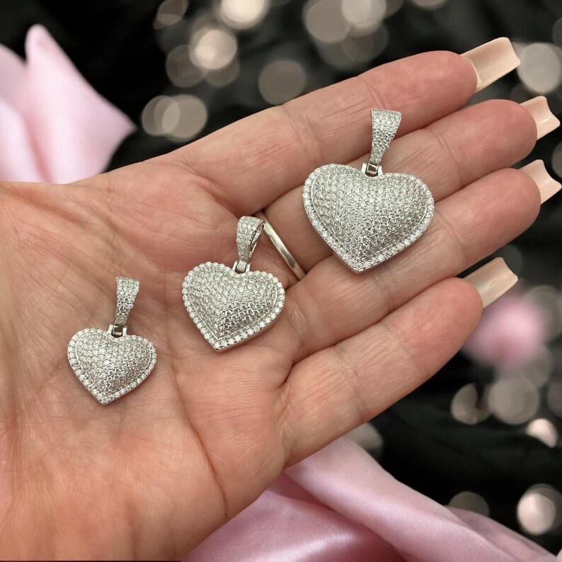 May include: Three silver heart pendants, each encrusted with small, sparkling stones. The pendants vary in size, with a bail at the top for a chain. The hearts are set against a blurred background with pink fabric.