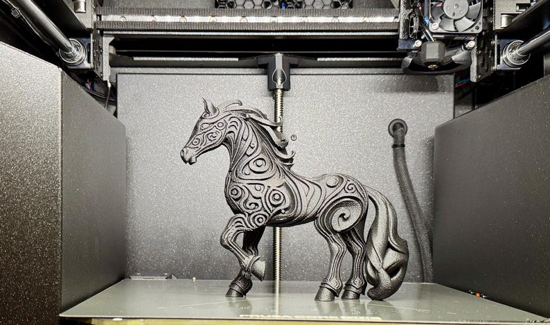 Puede incluir: Una figura decorativa de caballo negro con intrincados patrones de remolinos, dentro de una impresora 3D. El caballo tiene una melena y cola fluidas, y est&aacute; posado de forma din&aacute;mica. El fondo es gris oscuro.