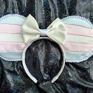 May include: White headband with two large ears, each with a white pom-pom trim and a pink, white, and blue striped fabric. A large white satin bow is centered on the headband.