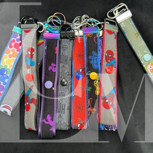 May include: A set of seven colorful keychains with different designs. The keychains are made of fabric and have a metal ring attached to them. The designs include a rainbow pattern, a spider-man character, a purple monster, a gray and black pattern, a red and black spiderweb pattern, a yellow and blue pattern, and a gray and black spider-man character.