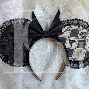 May include: Black Halloween-themed mouse ears headband. The ears feature a black bow, bat-shaped accents, and circular panels with a checkered pattern and spooky illustrations. The ears are trimmed with black lace.