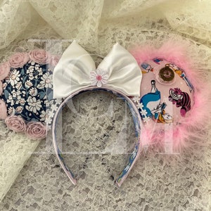 May include: A pair of white and pink Minnie Mouse ears with a white satin bow and a floral fabric design. One ear features a floral pattern and the other ear features a scene from Alice in Wonderland.