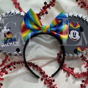 May include: A pair of gray Mickey Mouse ears with a rainbow bow. The ears are decorated with silver stars and feature Mickey Mouse and Minnie Mouse with rainbow accents. The headband is black.