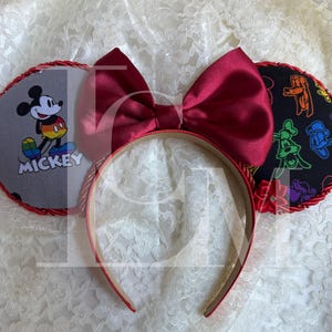 May include: A red headband with attached mouse ears and a large bow. One ear features Mickey Mouse with the word "Mickey". The other ear has colorful cartoon characters on a black background. The bow is a deep red.