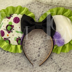 May include: Green and white Minnie Mouse ears with a black bow and purple flowers. The ears are decorated with a pattern of a green character with a sword and the text "PLUTO".