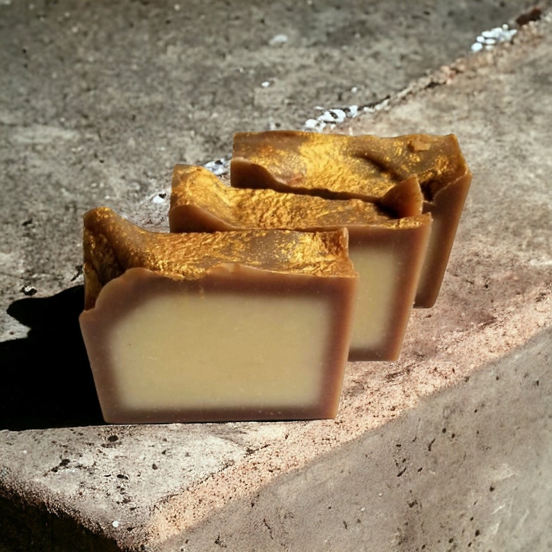 May include: Three bars of handmade soap with a brown base and gold flecks on top. The soap is stacked on a rough, gray surface.