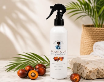 May include: White spray bottle of Island Magic Batana Oil hair growth mist. The bottle has a black spray nozzle and a label with a mermaid illustration and text. Several brown Batana nuts and a palm leaf are arranged around the bottle. The bottle contains 8 oz / 236ml.
