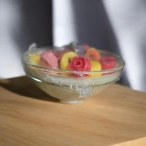 Puede incluir: Un bol de vidrio transparente lleno de fundidos de cera de colores, incluyendo amarillo, rosa y rojo. El bol está cubierto con film transparente.