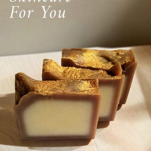 May include: Three bars of brown soap with gold flecks. The soap is stacked on a light wood surface. The text "Skincare For You" is written in white on a light gray background.
