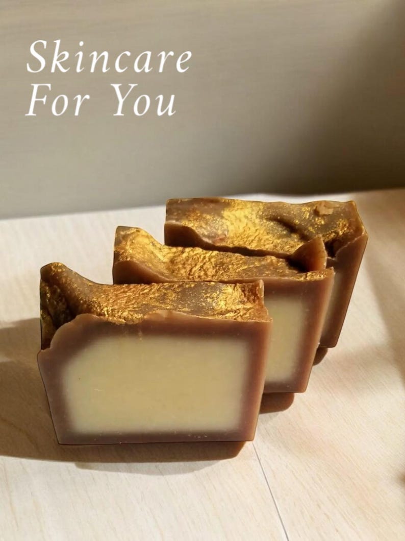May include: Three bars of brown soap with gold flecks. The soap is stacked on a light wood surface. The text "Skincare For You" is written in white on a light gray background.