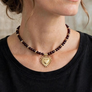 May include: A beaded necklace featuring dark red, faceted round beads interspersed with small, polished gold-tone metal spacers. The necklace includes a central gold-tone metal pendant shaped like a heart with radiating sunburst rays.
