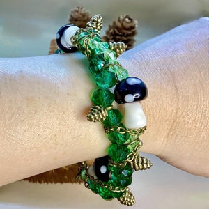 May include: A bracelet featuring vibrant green faceted crystal beads, small bronze-toned metal pinecone charms, and miniature black and white mushroom-shaped beads, all connected by delicate bronze-toned metal chains in a layered design.