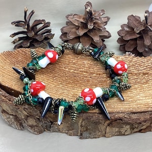 Puede incluir: Un brazalete con cuentas negras y verdes, abalorios de setas blancas y rojas y eslabones de cadena dorados. El brazalete tiene púas negras y está sobre una superficie de madera.