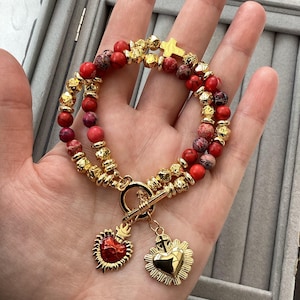 Può includere: Un bracciale a doppio filo con perline in pietra rossa e cremisi marmorizzato, alternate a perline in metallo dorato testurizzato. Il bracciale include un ciondolo a croce dorata e una chiusura a T con due pendenti a forma di cuore.