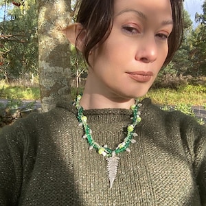 May include: A woman wearing a green sweater with a silver fern pendant necklace. The necklace is made of green and white beads.