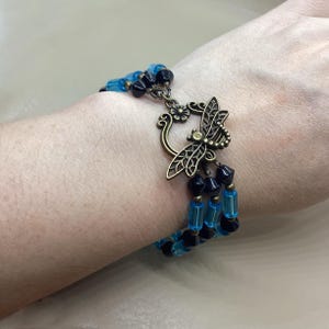 Puede incluir: Una pulsera de varias hebras con cuentas azules y negras. Tiene un cierre decorativo de libélula de color bronce. Las cuentas son una mezcla de formas redondas y rectangulares. La pulsera se lleva en la muñeca.