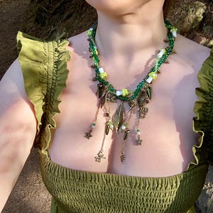 Puede incluir: Un collar de cuentas verdes y blancas con colgantes de hojas y flores doradas. El collar lo lleva una persona con un vestido verde.