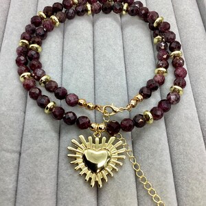 May include: A double-strand beaded necklace featuring faceted dark red garnet-coloured beads interspersed with small gold-toned metal spacers. A central gold-toned heart pendant with a sunburst-style frame hangs from the necklace chain.