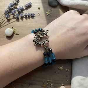 May include: A bronze dragonfly charm bracelet with blue and black beads. The bracelet features a decorative dragonfly clasp and dangling bead strands. The jewellery is displayed on a wooden surface with lavender and decorative charms.