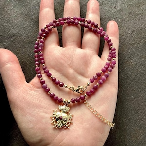 Puede incluir: Un colgante de corazón de oro con un centro de rubí y espinas, que cuelga de una doble hilera de cuentas de rubí rosa en una cadena de oro.