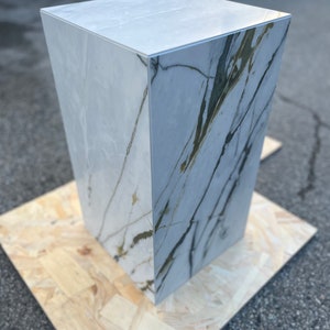 May include: A white marble pedestal with a light grey and green marbled pattern. The pedestal is sitting on a piece of plywood.