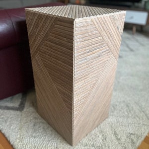 May include: A light brown wooden pedestal with a geometric pattern. The pedestal has a square top and a rectangular base.
