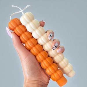 Puede incluir: Tres velas con forma de calabaza en tonos naranja y crema se sostienen en una mano. Las velas están dispuestas verticalmente, con una mecha blanca en la parte superior. El fondo es de color gris claro sólido.