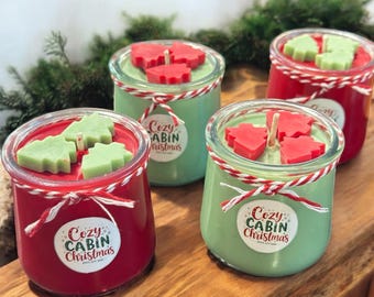 Puede incluir: Cuatro velas en tarros de vidrio rojos y verdes, decoradas para Navidad. Cada vela tiene un adorno de cera en forma de árbol y una etiqueta que dice "Cozy Cabin Christmas". Los tarros están atados con hilo rojo y blanco.