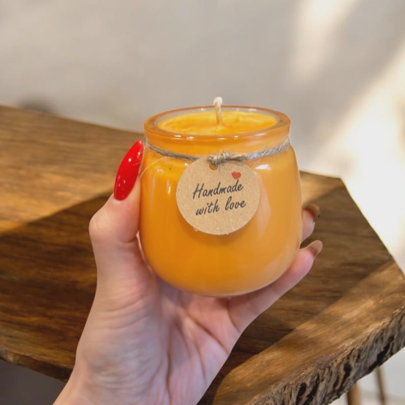 Puede incluir: Una vela naranja en un tarro de cristal. La vela tiene una mecha y est&aacute; adornada con una etiqueta redonda que dice "Handmade with love". El tarro est&aacute; atado con hilo.