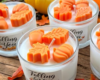 May include: Clear glass candles filled with white wax and topped with orange leaf, pumpkin, and acorn shapes. The candles have a label that says "Falling for You". Pumpkins and fall leaves are in the background.